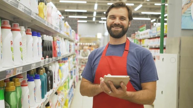 Handsome smiling supermarket employee with pc tablet standing among shelves