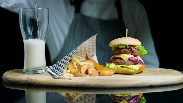 Chef serving set of tasty burger, french fries, pouring beer into glass on wooden tray