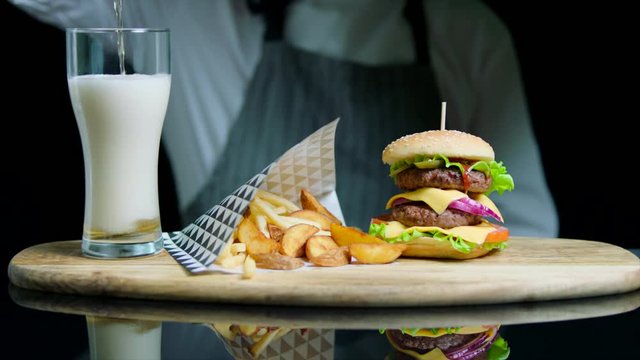 Chef serving set of tasty burger, french fries, pouring beer into glass on wooden tray