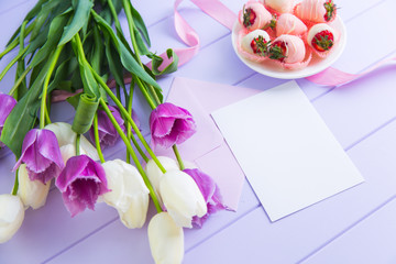 Clear sheet of paper with envelope, strawberry in chocolate, bouquet of white and purple tulips and pink ribbon on lilac table