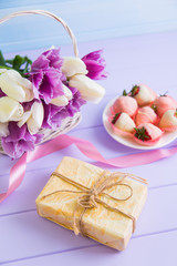 Gift box, strawberry in chocolate on plate, pink ribbon and bouquet of purple and white tulips in wicker basket on lilac table