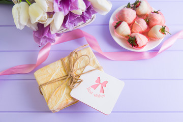 Obraz na płótnie Canvas Gift box, greeting card, strawberry in chocolate on plate, pink ribbon and bouquet of purple and white tulips on lilac table.