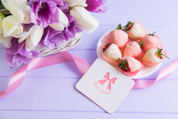 Greeting card, strawberry in chocolate on plate, pink ribbon and bouquet of purple and white tulips on lilac table.