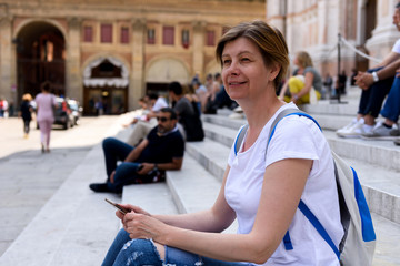 Middle-aged woman, tourist in Bologna, Italy. Sits on the steps of the Basilica of San Petronio in the central square of the city. Travel to Europe.