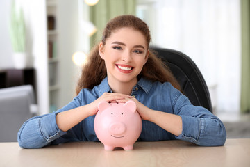 Obraz na płótnie Canvas Young woman holding piggy bank indoors. Money savings concept