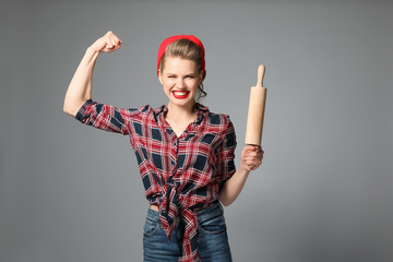 Strong young woman with rolling pin on grey background