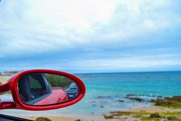Closeup of rearview mirror of a red car with defocused beach at the background