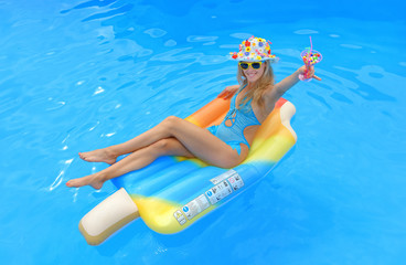 A young girl wearing a blue swim suit and a blue floral hat holds an imaginative cocktail drink in her hand. She  is lying on a swim ring in a swimming pool smiling at the camera.
