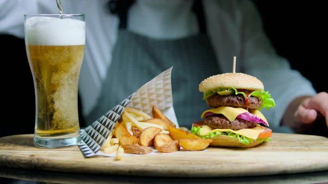 Chef is serving set of tasty burger, french fries, beer on the wooden tray on the black background
