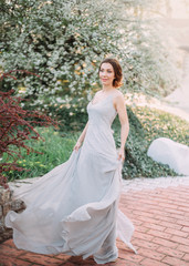 Red-haired girl in a modest, gray, silk dress in rustic style, which flutters in the wind. Portrait of the bride against the background of a flowering tree. Accurate, collected hair. Art photo