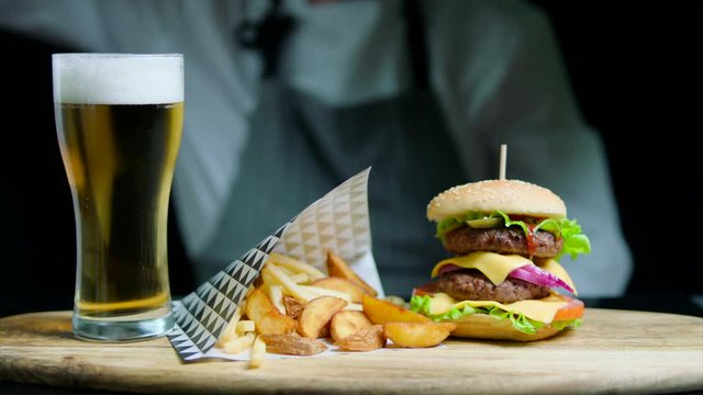 Set of tasty burger, french fries, beer on the wooden tray. Chef dancing on the background