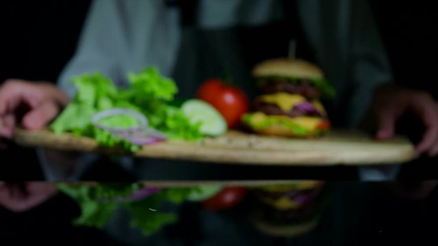 Fresh vegetables and tasty burger with cheese and beef cutlets. Chef on the background out of focus