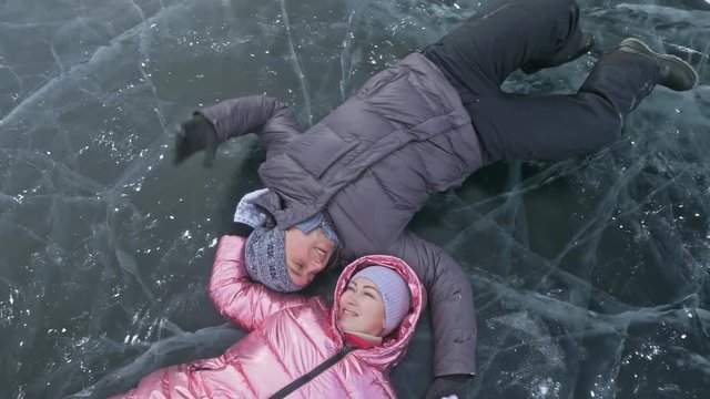 Young couple has fun during winter walk against background of ice of frozen lake. Lovers lie on clear ice with cracks, have fun, kiss and hug. View from above. Young happy people stroll through snow