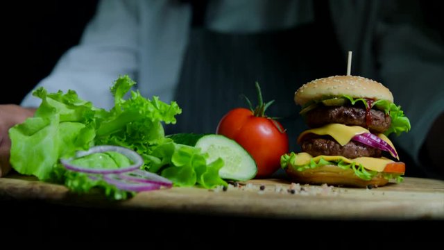 Fresh vegetables and tasty burger with cheese and beef cutlets. Chef on the background out of focus
