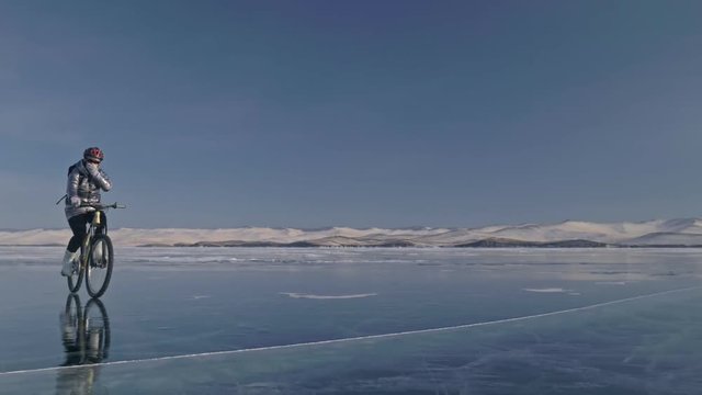 Woman is riding bicycle on the ice. The girl is dressed in a silvery down jacket, cycling backpack and helmet. Ice of the frozen Lake Baikal. The tires on the bicycle are covered with special spikes