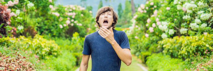 Young man sneeze in the park against the background of a flowering tree. Allergy to pollen concept...
