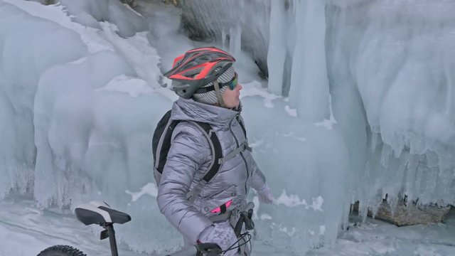 Woman is walking beside bicycle near the ice grotto. The rock with ice caves and icicles is very beautiful. The girl is dressed in silvery down jacket, cycling backpack and helmet. The tires on
