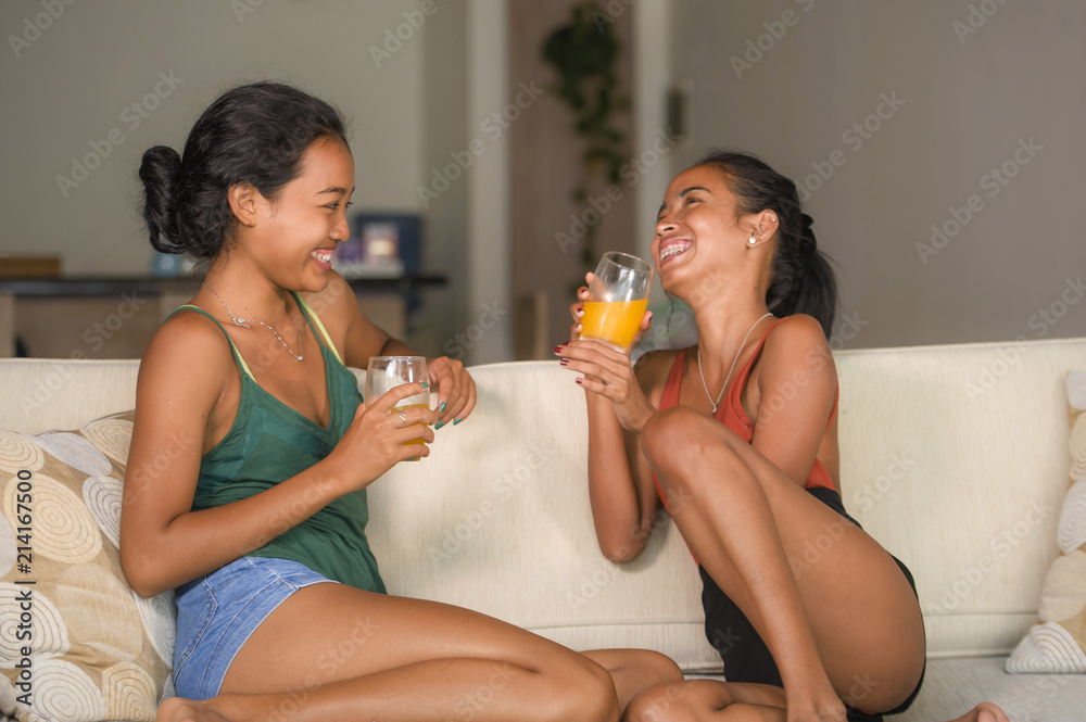 Wall mural lifestyle portrait of two young happy and relaxed Asian girlfriends having fun talking laughing and gossiping at home sofa couch sharing complicity together