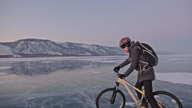 Man is riding a bicycle on ice. The cyclist is dressed in a gray down jacket, backpack and helmet. Ice of the frozen Lake Baikal. The tires on the bicycle are covered with special spikes. The traveler