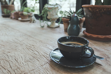 A black cup of hot coffee and a small tree pot on vintage wooden table in cafe