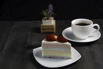 Rainbow crape cake with fruit fresh strawberry on white plate with a cup of hot tea on dark wooden table, isolated black background.