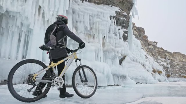 Man is walking beside bicycle near the ice grotto. The rock with ice caves and icicles is very beautiful. The cyclist is dressed in gray down jacket, cycling backpack and helmet. The tires on covered