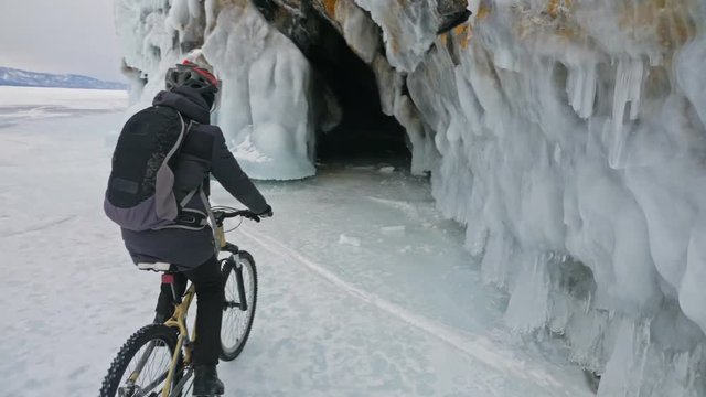 Man is riding bicycle near the ice grotto. The rock with ice caves and icicles is very beautiful. The cyclist is dressed in gray down jacket, cycling backpack and helmet. The tires on covered with