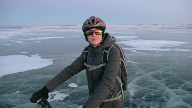 Man is stand a bicycle on ice. He looks at the beautiful sunset. The cyclist is dressed in a gray down jacket, backpack and helmet. Ice of the frozen Lake Baikal. The tires on the bicycle are covered