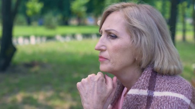 Thoughtful aged lady covered with plaid blanket sitting on bench in garden