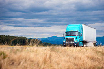 Blue modern big rig semi truck transport semi trailer on scenic road with grass forest mountain and cloudy sky © vit