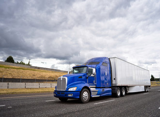 Fototapeta na wymiar Classic blue big rig semi truck with dry van semi trailer going on wide highway with stormy sky