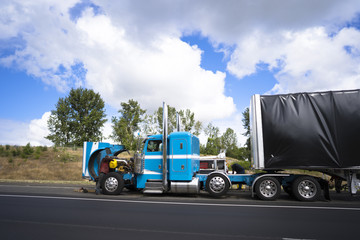 Truck drivers repairing big rig semi truck with open hood right on the road shoulder