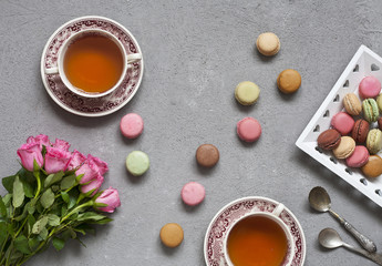 Close-up of a bouquet of pink roses, two cups of tea and fresh colorful  macaroons. Top view of a gray concrete background