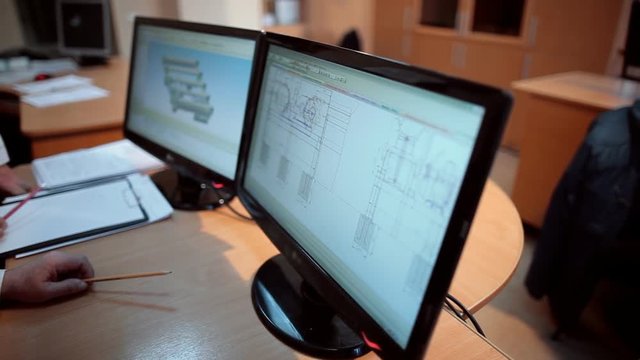 In the office, 2 engineers review and discuss the details in the diagrams of equipment, over which they work. By pencils, they accurately indicate the place to look at