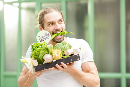 Portrait of a happy vegetarian man holding box full of fresh raw vegetables biting pepper outdoors on the green background