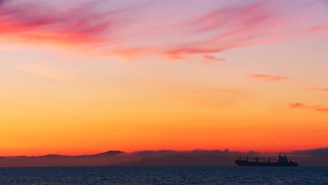 A huge cargo ship stands on a roadstead at sea in the yellow and red rays of the evening sun against the background of distant mountains
