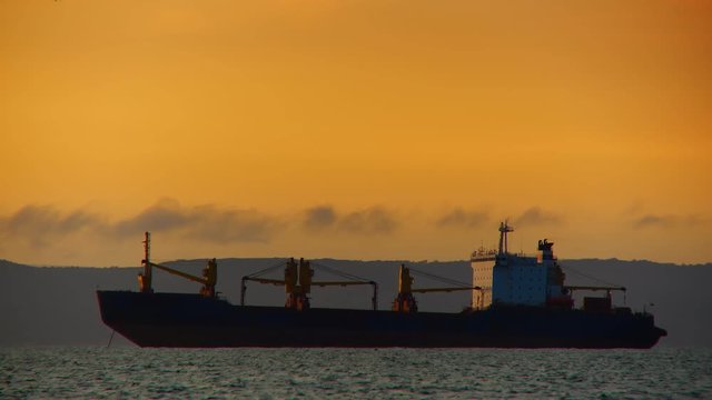 A huge cargo ship stands on a roadstead at sea in the yellow rays of the evening sun
