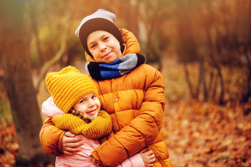 autumn portrait of happy kids playing and hugs outdoor in park. Smiling brother and sister walking in sunny day, wearing warm knitted hats and scarfs