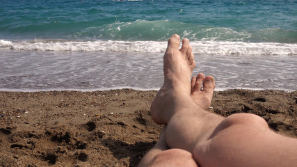 Point of view, first person view. The legs of a man against the background of the sea. A man is lying on the beach, looking at his feet and the sea.
