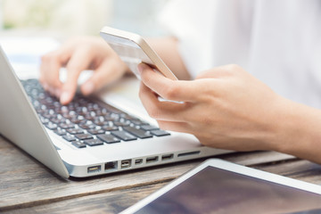 Fototapeta premium Woman one hand holding a phone while the other hand typing on laptop keyboard, outdoor in the garden