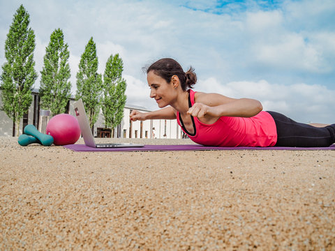 fit and sporty woman doing fitness workout online infront of a laptop on a yoga mat outside in the park 