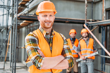 smiling builder standing at construction site with crossed arms while his colleagues standing...