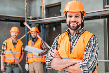 handsome smiling builder standing at construction site with crossed arms while his colleagues standing blurred on background - obrazy, fototapety, plakaty