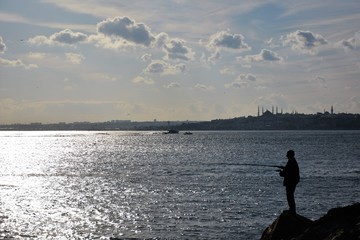 Fototapeta na wymiar A man is fishing on the rocks by the city silhouette