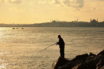 A man is fishing on the rocks by the city silhouette