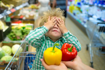 Choice blindly. Hide and seek. Shopping with children. Pepper. Red or yellow pepper. Vegetables in store. Child in shopping cart. No appetite. Vegetable diet. Funny baby dont want eat healthy food - 214207944