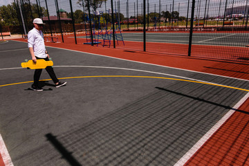 Modern man in stylish wear walks at sport playground with yellow skateboard in hand