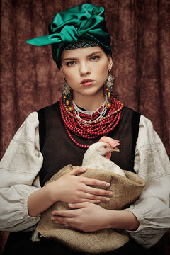 Portrait of a young pretty woman sitting in national rustic Slavic costume, wearing green headwrap, red necklace and long earrings, holding sack with white hen, looking at camera over brown background