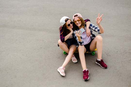 Two young happy girl friends in hipster outfit sitting together on longboard and making selfie on phone