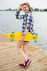 Young cheerful girl in hipster outfit holding yellow longboard in his hand and walking on a wooden pier